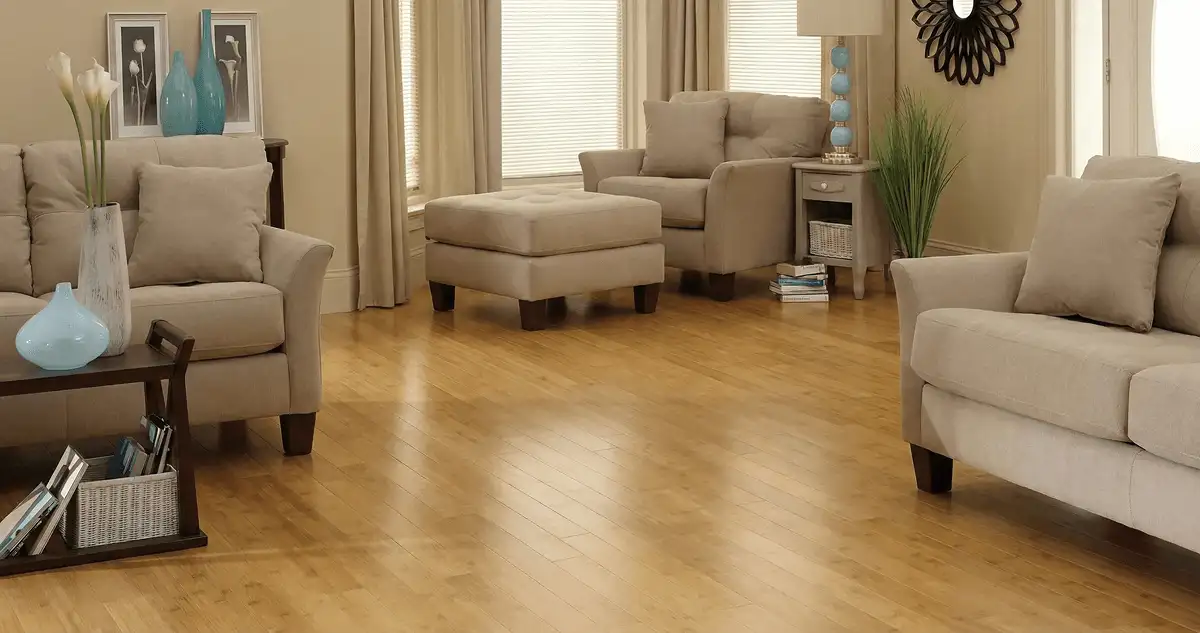 A living room with professionally installed bamboo flooring, beige sofas, a matching ottoman, a side table with a blue lamp, vases, framed pictures, and large windows with blinds and curtains in Tuscaloosa, AL